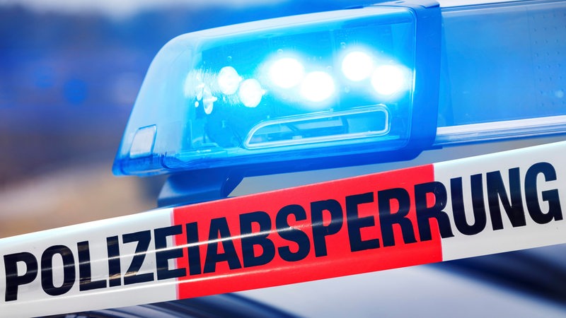 Ein Absperrband der Polizei mit der Aufschrift "Polizeiabsperrung". Im Hintergrund ist ein Blaulicht zu sehen.