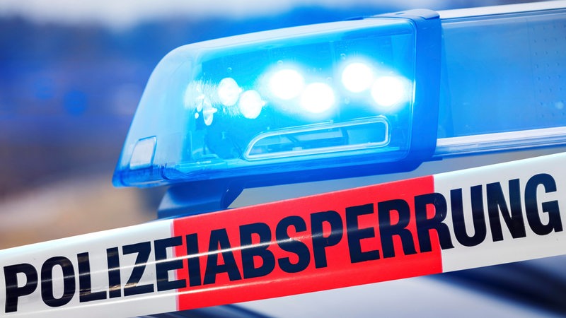 Ein Absperrband der Polizei mit der Aufschrift "Polizeiabsperrung". Im Hintergrund ist das Blaulicht eines Polizeiautos zu sehen.