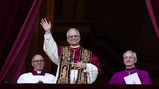 Der neue Papst Leo XIV winkt von einem Balkon, neben ihm sitzen zwei Männer.