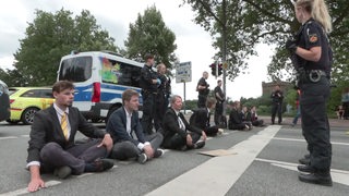 Mehrere junge Menschen in Anzügen sitzen im Schneidersitzt auf der Fahrbahn.