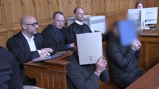 Zwei Männer halten sich Aktenordner vors Gesicht während sie auf der Anklagebank sitzen.