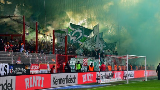 In einem Fußballstadion brennen Zuschauer Feuerwerkskörper ab. Es steigen grüne Rauchschwaden auf.