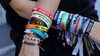 Eine Frau zeigt ihre Arme mit verschiedene queere Armbänder und Bändchen (Symbolbild)