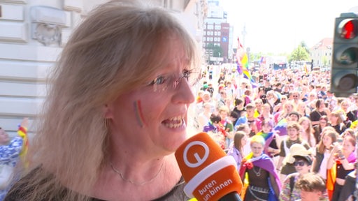 Eine Frau wird interviewt, dahinter Christopher Street Day Umzug