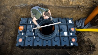 Ein Mann verbaut im Garten neben seinem Haus eine Wasserzisterne, um seinen Garten mit Regenwasser bewaessern zu koennen.