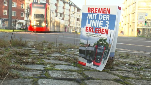 Ein Reiseführer steht auf dem Steinboden an der Bahn-Haltestelle. Im Hintergrund kommt die Bahn der Linie 3 angefahren.