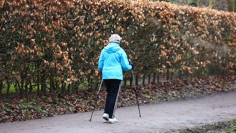 Eine Frau läuft auf einem Weg neben einer Hecke mit braunen Bättern. Sie trägt eine hellblaue Jacke, eine schwarze Hose und läuft mit zwei Nordic-Walking-Stöcken-