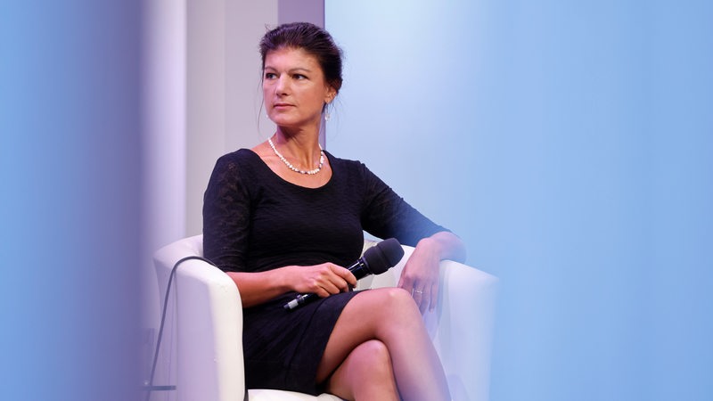 Sahra Wagenknecht sitzt in einem weißen Sessel und hält ein Mikro in der Hand.