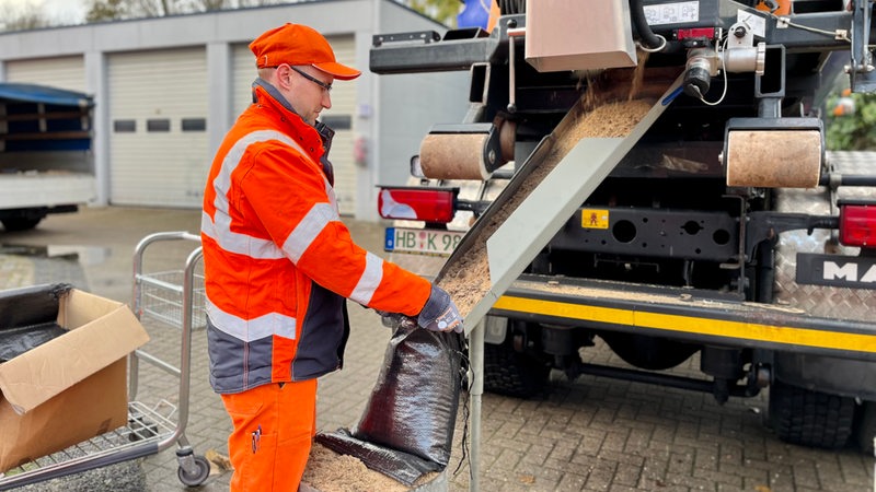 Ein Mann in orangefarbener Kleidung steht mit einem Sack in der Hand hinter einem Streuwagen.