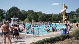 Blick auf Freibad bei Sonnenschein mit vielen Menschen im und am Wasser. 