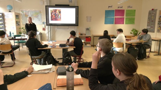 Mehrere Schüler in einer Schulklasse gucken an eine digitale Tafel und zu ihrer Lehrerin im Klassenraum 