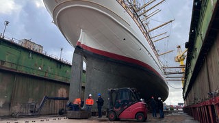 Menschen und ein Gabelstapler stehen unter einem Schiff im Dock.