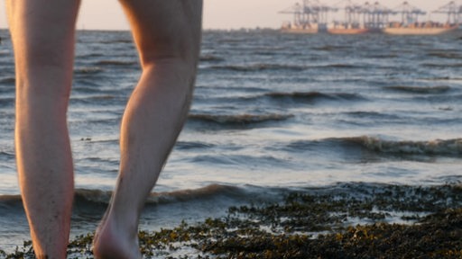 An einem Strand sind im Vordergrund zwei nackte Beine zu sehen.