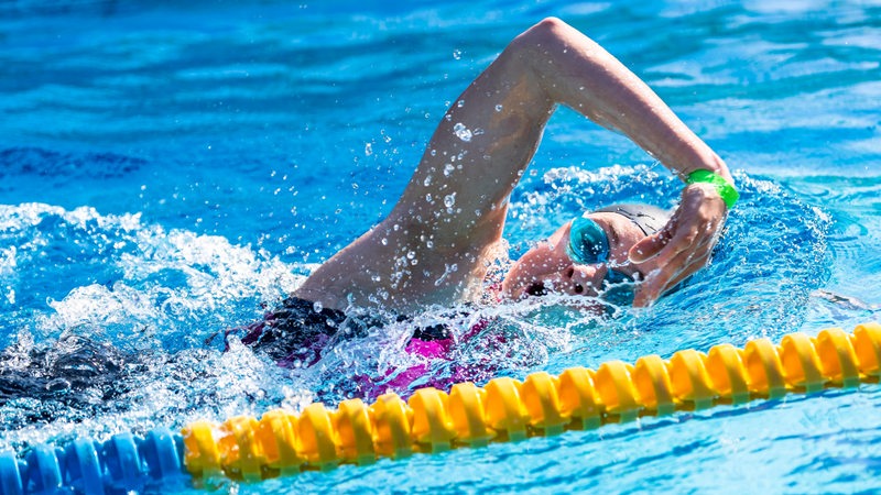 Eine junge Triathletin schwimmt im Kraulstil im Schwimmbad, mit hohem Ellbogen holt sie den Arm zurück nach vorne und atmet dabei ein.