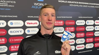 Schwimm-Olympiasieger Florian Wellbrock steht vor einer Werbewand beim Interview und hält seine Silbermedaille der Kurzbahn-WM in die Kamera.