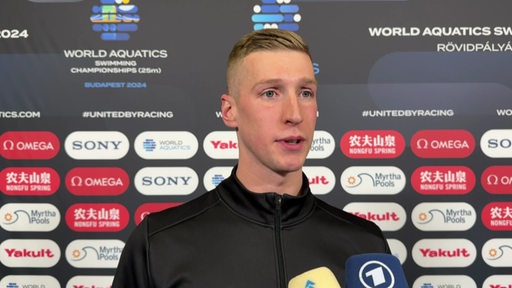 Schwimmstar Florian Wellbrock steht nach seinem Gewinn der Silbermedaille bei der Kurzbahn-WM vor einer Werbewand beim Interview.