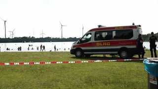 An einem Badesee ist ein Teil durch Flatterband abgesperrt, daneben steht ein Einsatzfahrzeug der DLRG