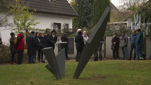 Eine graue Skulptur steckt in einer Rasenfläche, im Hintergrund einige Menschen