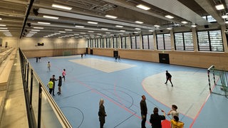 Ein Blick in eine Schulsporthalle: Kinder laufen durch die Halle und stehtn teilweise am Rand