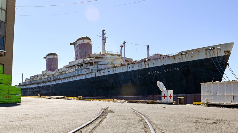 Ein marodes, großes blau-weißes Schiff mit zwei Schornsteinen liegt an einem Pier.