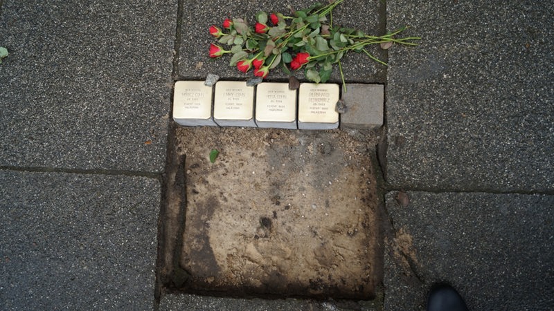 Kleine helle Steine liegen nebeneinander in einem Loch im Pflaster. Daneben liegen Rosen.