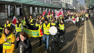 Streikende Menschen mit Transparenten und Fahnen ziehen am Bahnhof vorbei