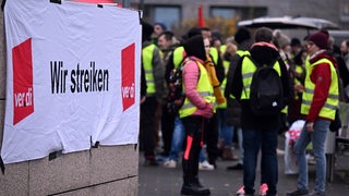 Hinter einem Transparent mit der Aufschrift: "Wir streiken/Verdi", stehen in gelben Warnwesten streikende Menschen.