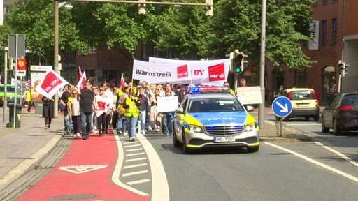 Auf einer Straße ist ein Warnstreik mit vielen Menschen zu sehen. Vor der Gruppe fährt ein Polizeiwagen.