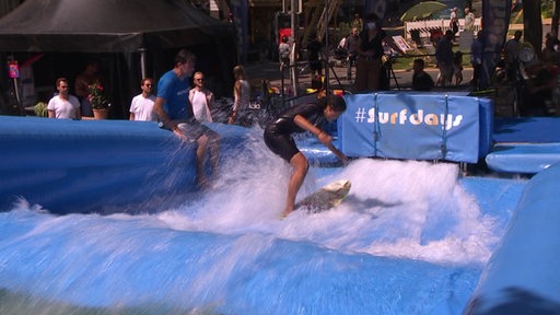 Eine Surferin in einem blauen Schwimmbecken auf ihrem Brett. Der Surflehrer sitzt auf dem Schwimmbeckenrand. Im Hintergrund Zuschauer.