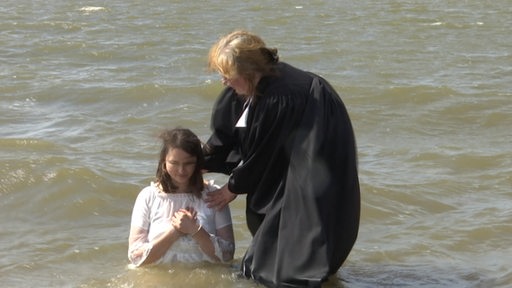 Eine Pfarrerin steht neben einer im Wasser knienden Frau.