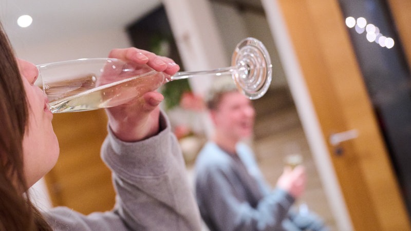 Ein Teenager trinkt ein Glas Sekt, während ein weiterer Jugendlicher im Hintergrund ebenfalls Alkohol in der Hand hat. (Symbolbild)