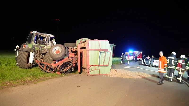 Der Anhänger eines Traktors liegt umgekippt auf einer Landstraße. Der Traktor steht neben der Fahrbahn. Feuerwehrleute stehen auf der Straße. Ein stark beschädigtes Auto steht auf der Straße.