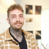 Stark tätowierter Mann, etwa Mitte 30, guckt vor Ausstellungsbildern in die Kamera
