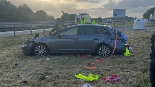 Ein kaputtes Auto steht auf einem Grasstück neben einer Straße, Gegenstände liegen verteilt auf dem Boden.