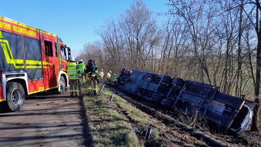 Links steht ein Feuerwehrauto. Daneben stehen Feuerwehrleute. Rechts liegt ein Lkw auf der Seite im Gras.