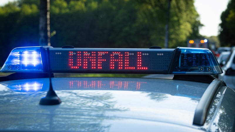 Ein Einsatzfahrzeug der Polizei steht mit Blaulicht und dem Schriftzug Unfall im Display an einem Unfallort.