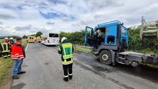 Feuerwehrleute und ein Mann in Warnjacke stehen an einer Straße neben einem Kipplaster und ein Schulbus.