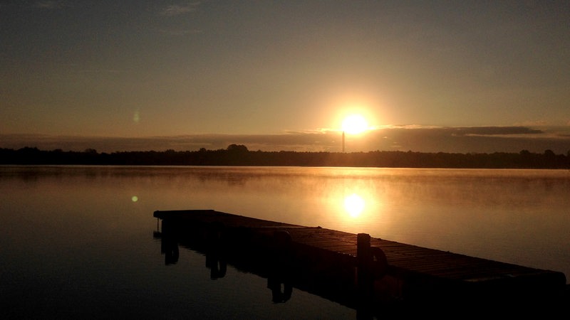 Die Sonne geht über einem See unter. Im Vordergrund ist ein Steg zu sehen.