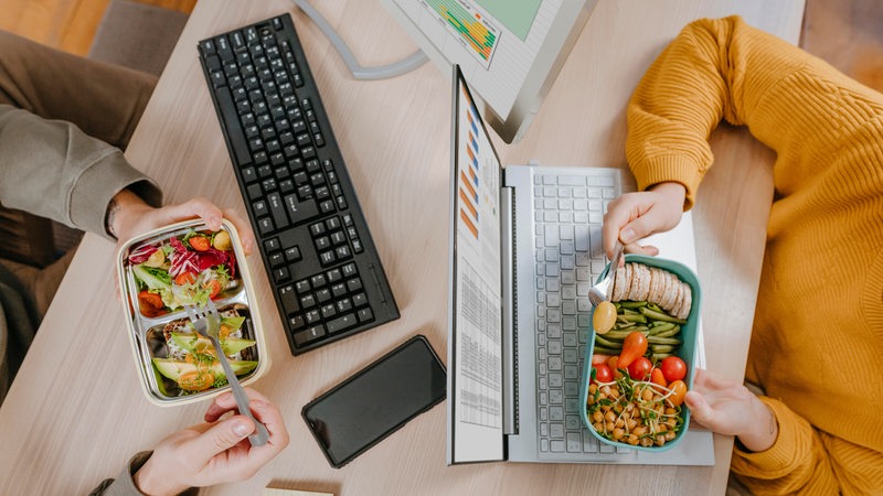 2 Leute sitzen sich an Laptops gegenüber und essen veganes Frühstück aus ihren Boxen.