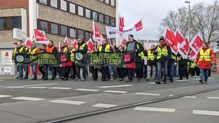 Menschen in Warnwesten und mit Verdi-Fahnen laufen über eine Straße.