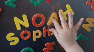 Eine Kinderhand berührt Magnetbuchstaben, die die Vornamen Noah und Sophie bilden. 