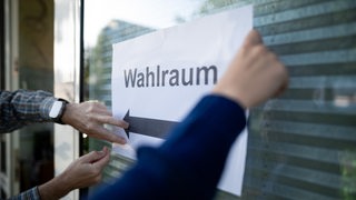 Hände kleben einen Zettel an eine Scheibe. Darauf steht "Wahlraum".