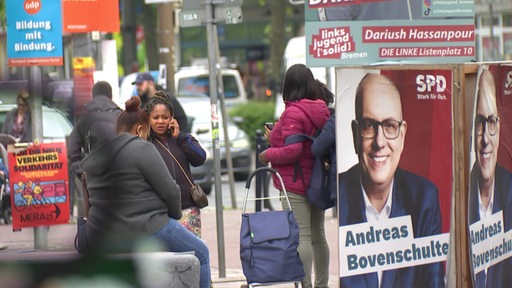 Mehrere Menschen stehen neben den Wahlplakaten auf der Straße. 