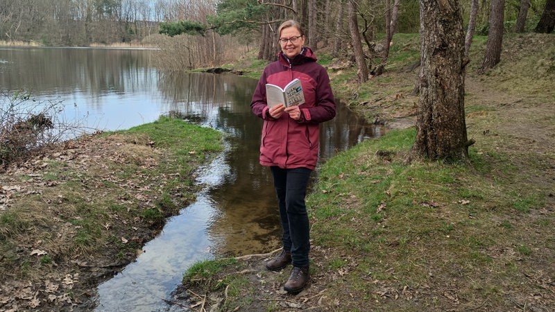 Eine Frau steht mit einem Wanderführer an einem See.