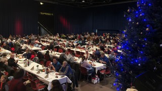 Zahlreiche Menschen sitzen an langen Tafeln neben einem beleuchteten Weihnachtsbaum.