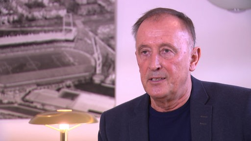 Dieter Burdenski bei einem Interview in seinem Büro mit einem Bild des Weserstadions hinter seinem Schreibtisch.