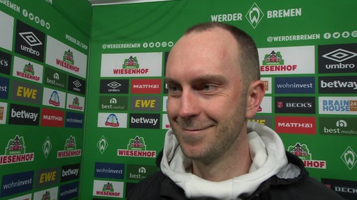 Werder-Coach Ole Werner steht lächelnd vor einer grünen Werbewand beim Interview nach dem Bayern-Spiel.