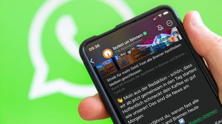 Eine Hand hält ein Smartphone mit dem WhatsApp-Channel von buten un binnen. Im Hintergrund das WhatsApp Logo