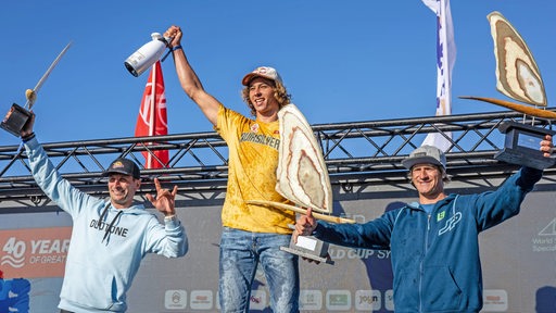 Windsurfer Lennart Neubauer steht auf einem Podest. Links und rechts von ihm stehen der Zweit- und Drittplatzierte.