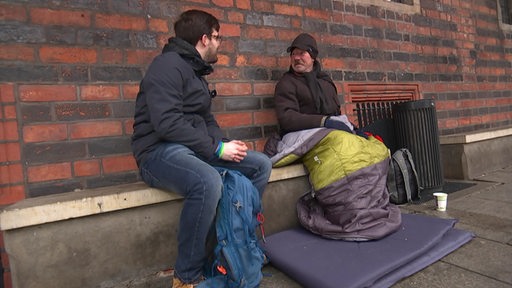 Eine Person spricht mit einer Wohnungslosen Person, die in einem Schlafsack sitzt